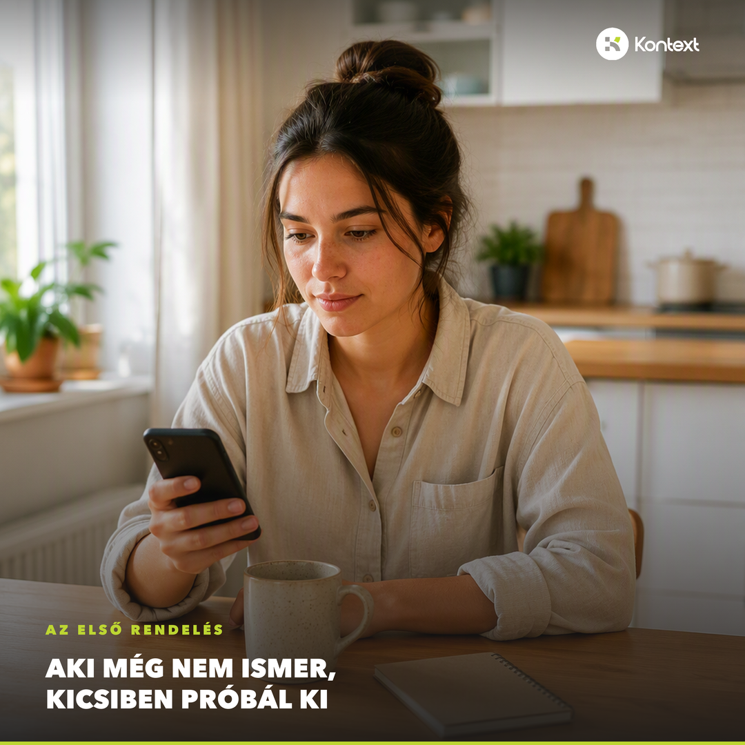 Nő a konyhaasztalnál a telefonját nézi, mellette bögre és jegyzetfüzet: az első, óvatos rendelés pillanata.